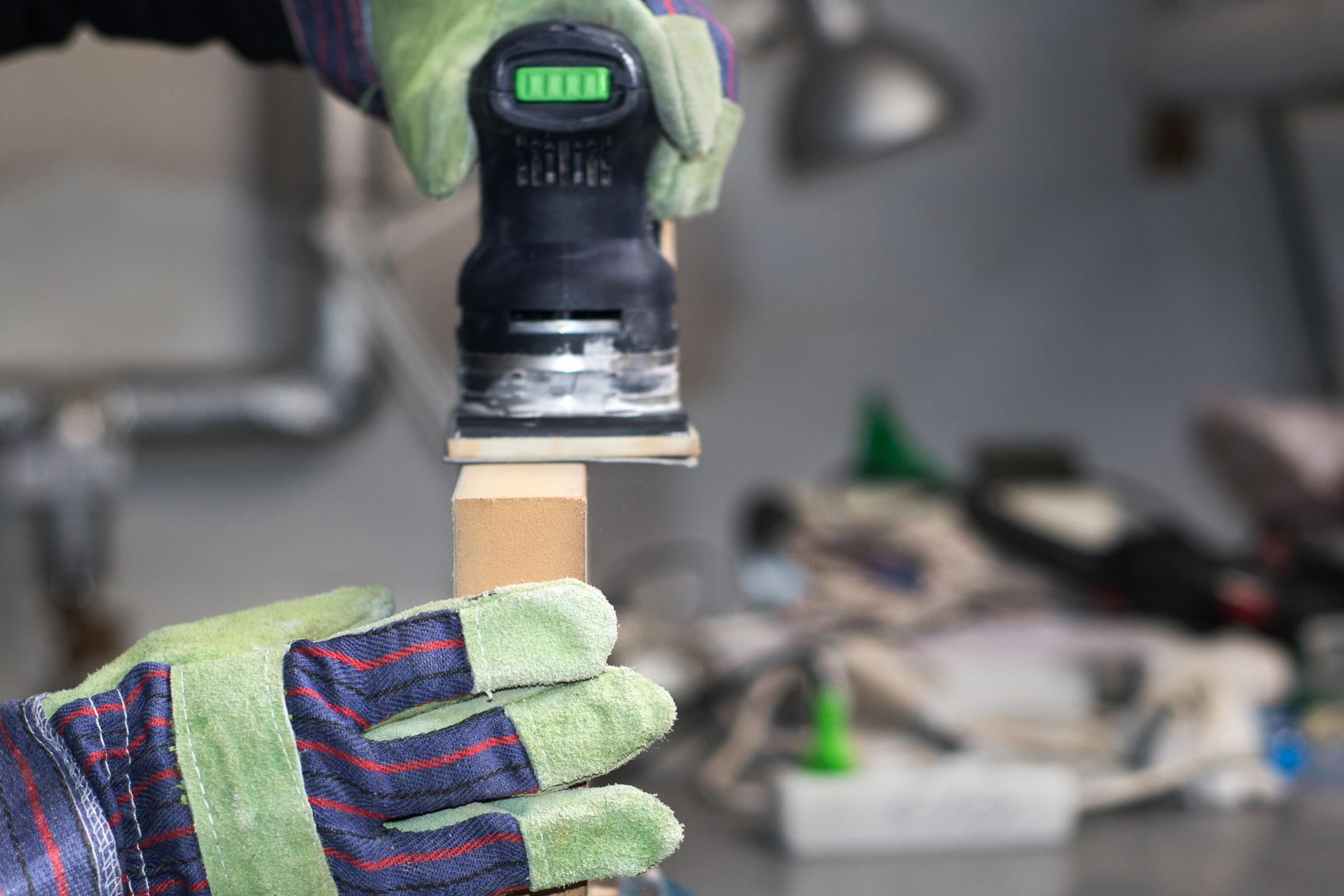 Nahaufnahme in einer Holzwerkstatt: Zwei Hände in Arbeitshandschuhen halten eine Schleifmaschine und bearbeiten damit ein Stück Holz