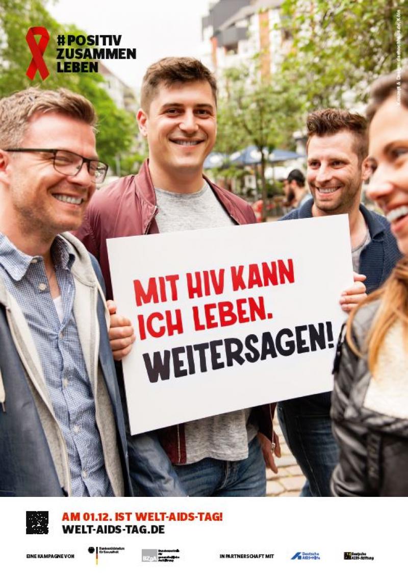 Mann im Freundeskreis hält ein Schild hoch. Aufschrift: "Mit HIV kann ich leben. Weitersagen!"