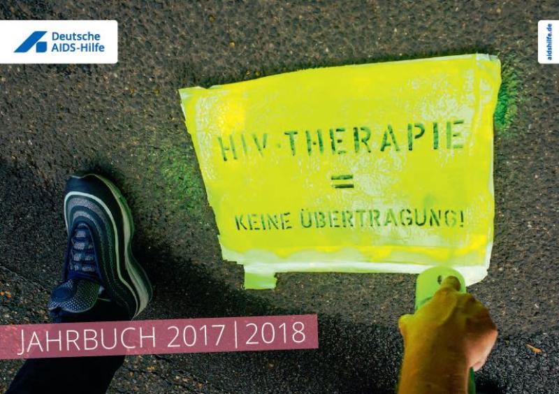 Asphalt, Füße und Hand einer Person, welche mit einer gelben Farbdose und einer Schablone den Text "HIV-Therapie = keine *Übrtragung" auf den Boden sprüht