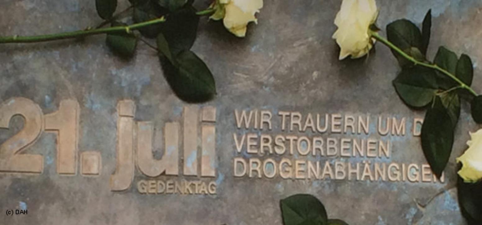 Zu sehen ist ein Gedenkstein mit der Aufschrift: 21. Juli, Gedenktag, wir trauern um die verstorbenen Drogenabhängigen. Frische weiße Rosen umrahmen die Aufschrift.