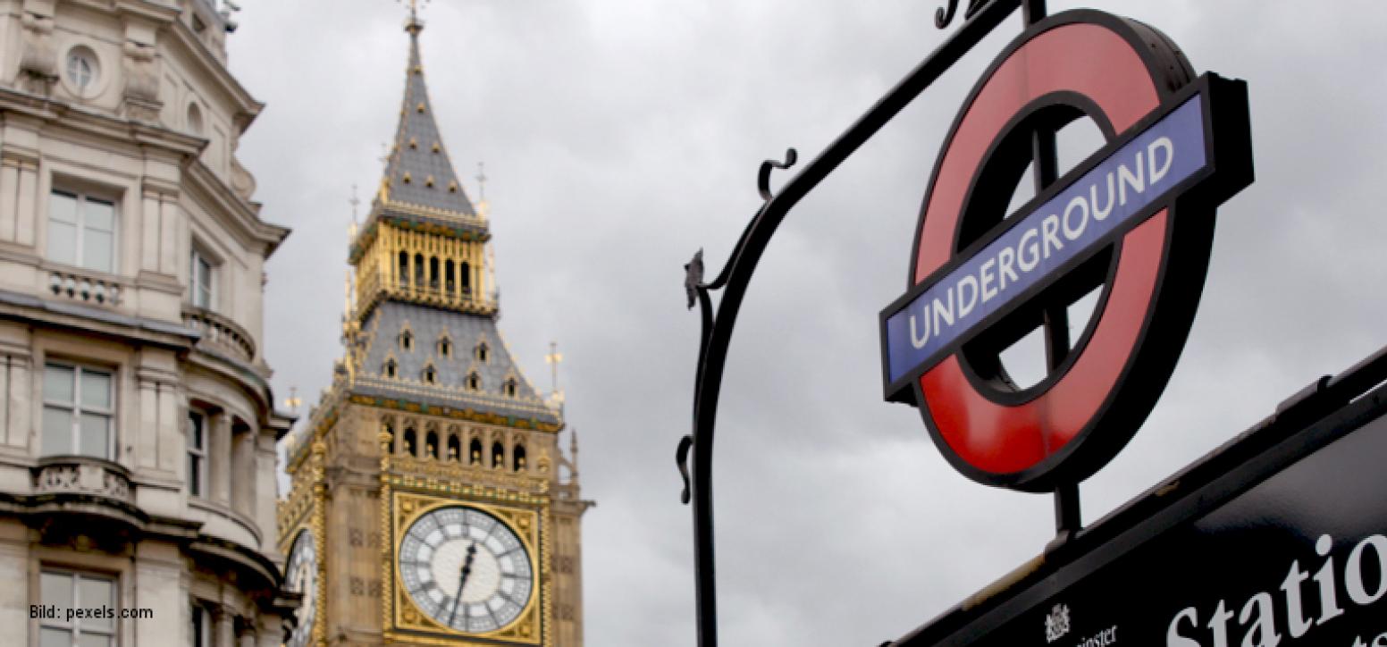 Im Vordergrund ist das Schild zu einem Eingang der Londoner U-Bahn mit der Beschriftung "Underground" zu sehen. Im Hintergrund sieht man den oberen Teil des Turms der Houses of Parliament mit der Uhr von Big Ben.