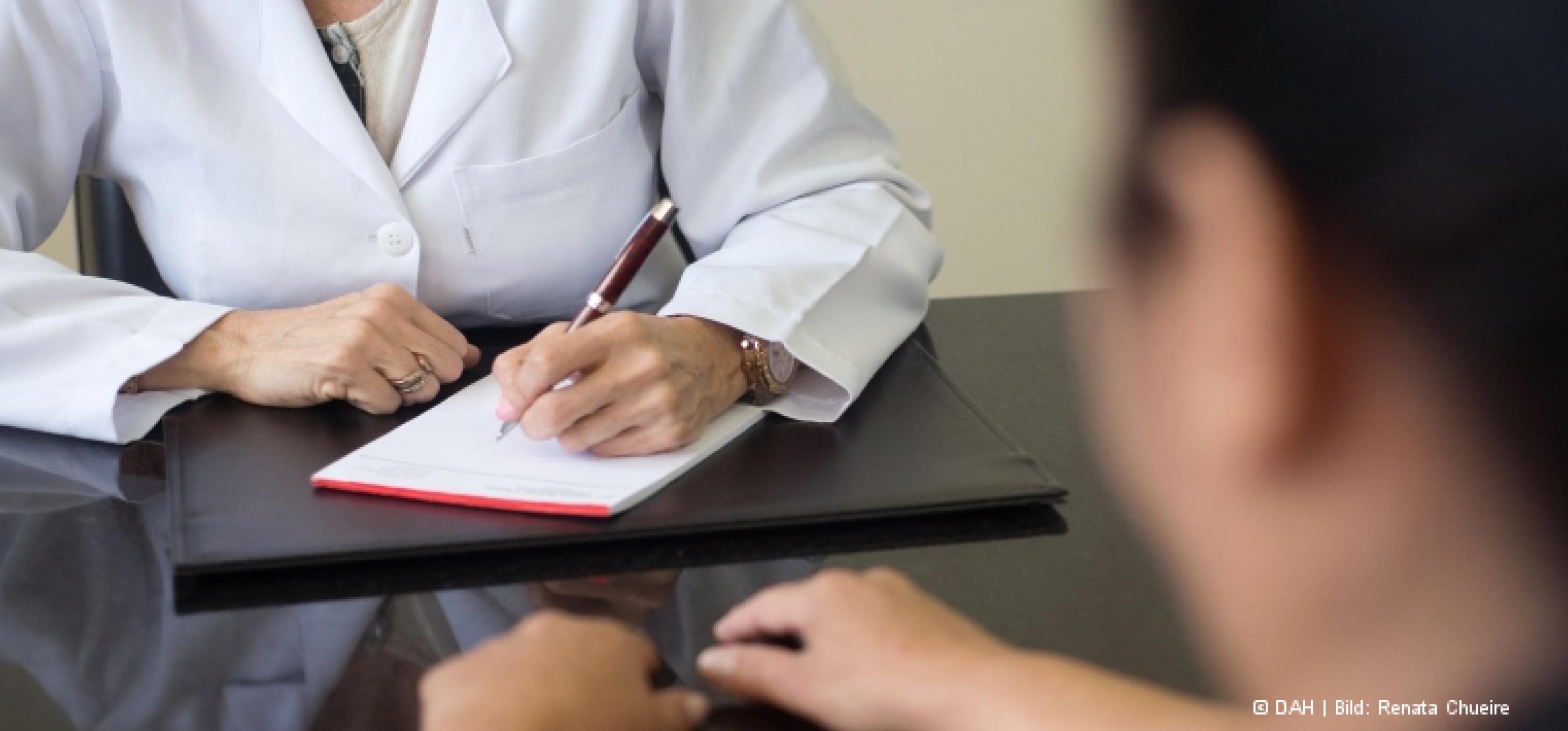 Eine Ärztin im weißen Kittel sitzt an ihrem Schreibtisch und macht sich Notizen auf einem Block, ihr gegenüber sitzt ein Patient