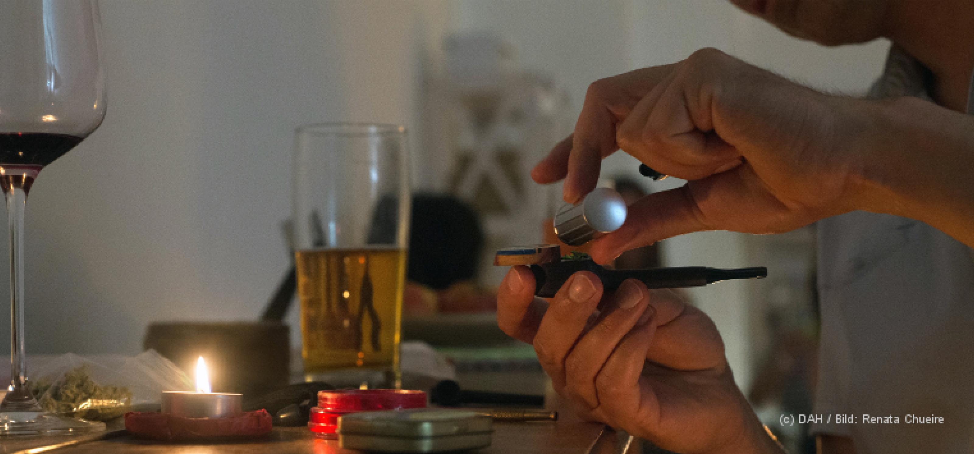 Auf einem Tisch befinden sich verschiedene Drogen: Ein Glas Bier, ein Glas Wein, eine Tüte mit Cannabis, zudem ein Sniefröhrchen, ein brennendes Teelicht und ein Mörser. Davor sitzt eine Person, die sich etwas in eine Pfeife füllt.