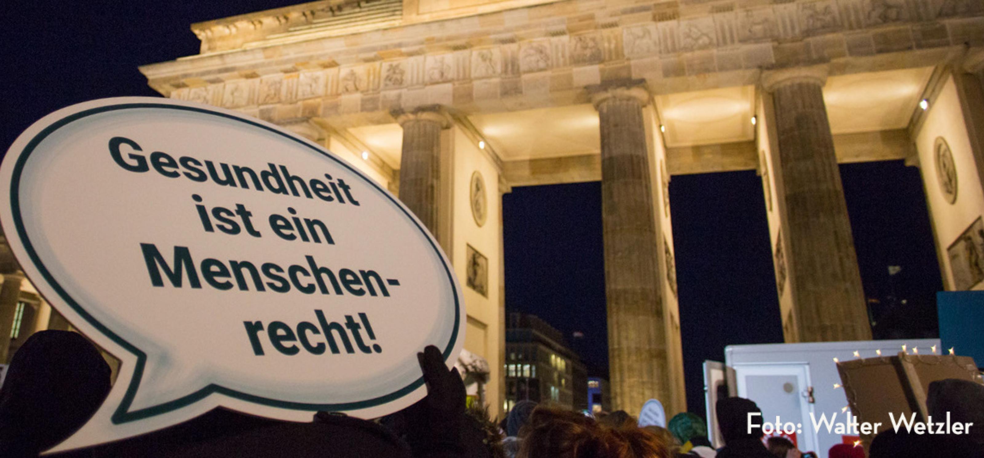 Eine Demonstration am Brandenburger Tor in Berlin, im Vordergrund ein Schild mit der Aufschrift "Gesundheit ist ein Menschenrecht"