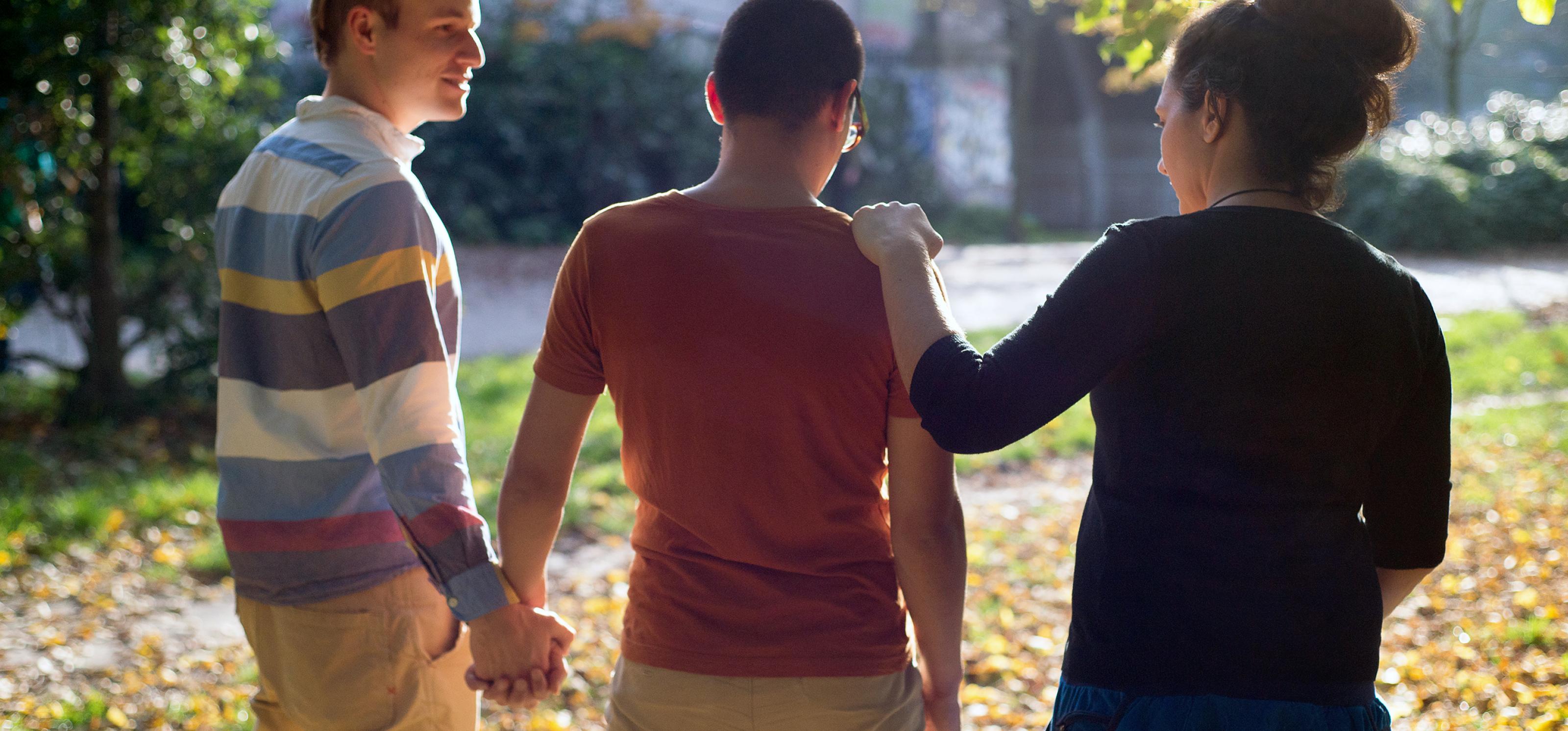 Eine Gruppe von drei Personen geht durch einen herbstlichen Park: Zwei Männer halten sich an den Händen, eine Frau legt ihre Hand auf die Schulter des einen Mannes