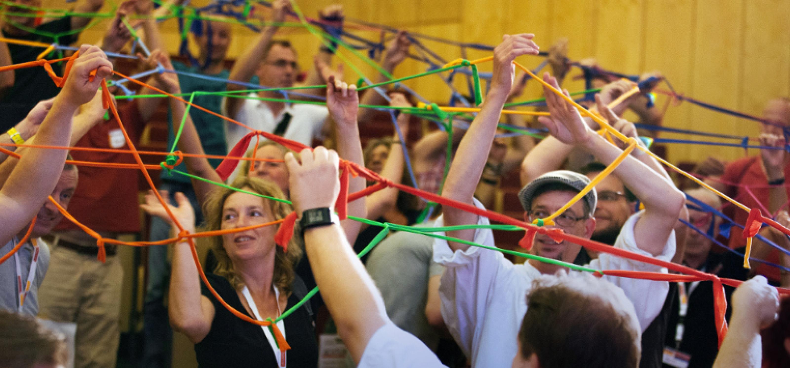 Viele Menschen in einem Konferenzsaal halten an ausgestreckten Armen ein Netz aus zusammengeknüpften bunten Bändern in die Luft